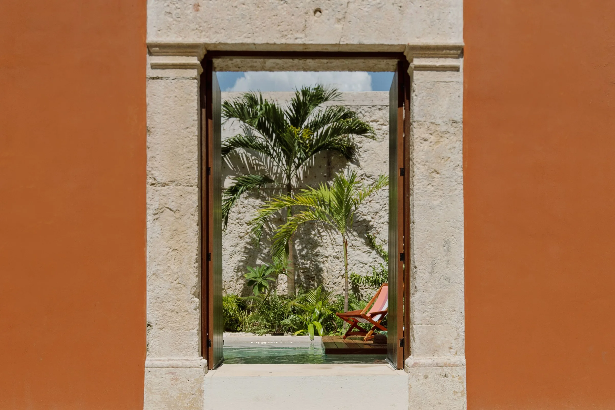 Puerta abierta en la fachada terracota de Casa Ex-Templo, con palmeras y la alberca del patio al fondo, en la ciudad amurallada de Campeche. Casonas MX